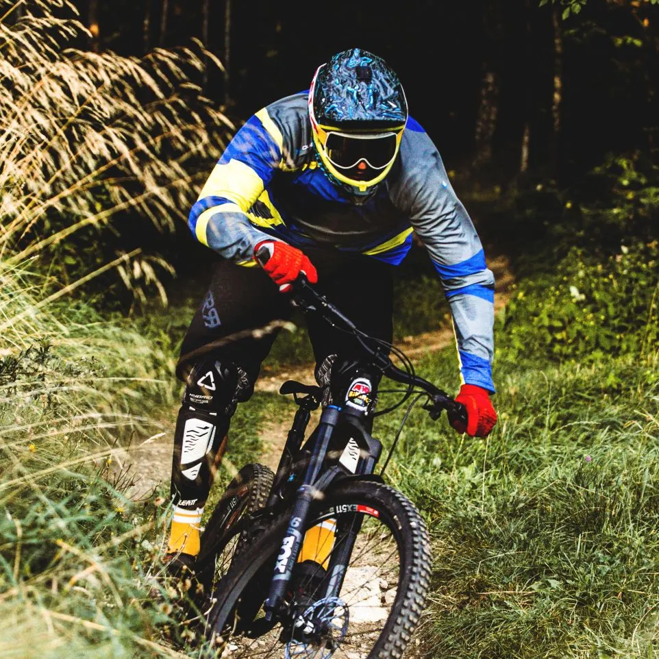 Ein Mountainbiker in einem farbenfrohen, langärmeligen Trikot mit blauen, gelben und grauen Akzenten fährt auf einem Waldweg.