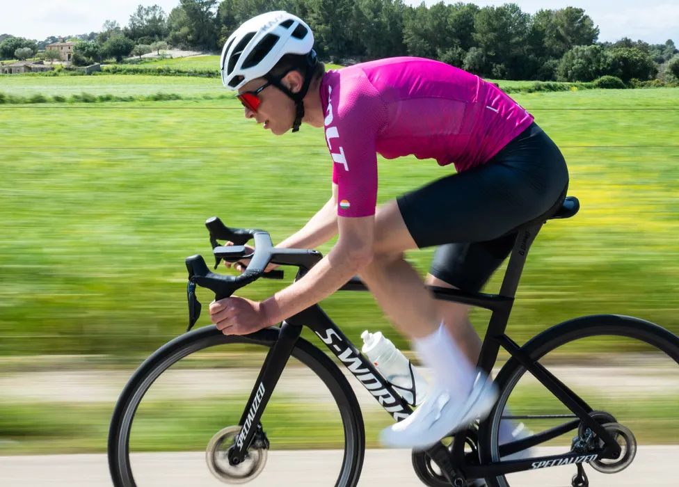 Ein Radfahrer in einem pinken Trikot und schwarzen Shorts fährt auf einem Rennrad auf einer Straße durch eine grüne Landschaft, wobei die Bewegungsunschärfe Geschwindigkeit vermittelt.