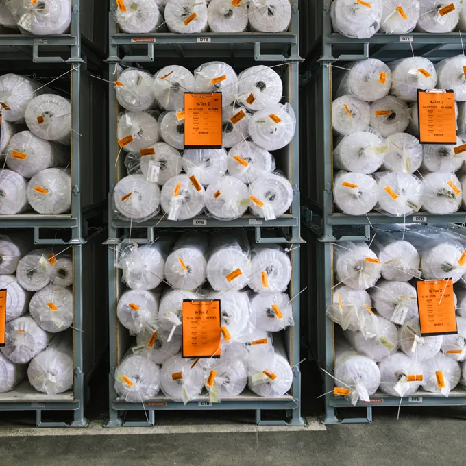 Ein Lager mit Regalen, die mit großen Rollen weißer Stoffe gefüllt sind, die in Plastik verpackt sind und orangefarbene Etiketten tragen.