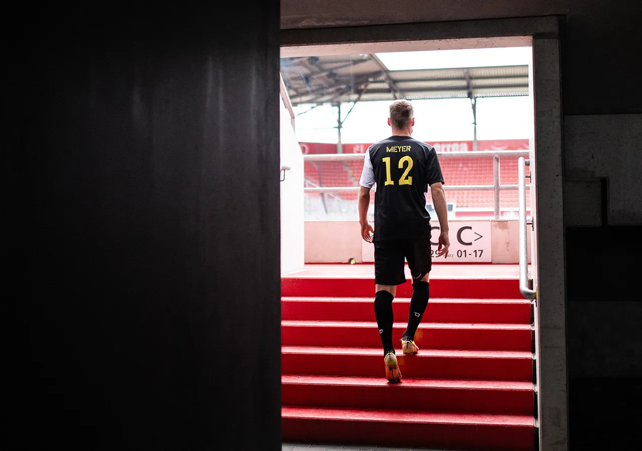 Ein Fußballspieler mit der Rückennummer 12 und dem Namen Meyer auf dem Trikot steigt eine rote Treppe in ein Stadion hinauf.